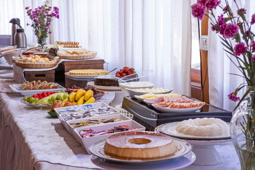 Mesa de buffet com sobremesas variadas, frutas e pratos, disposta ao lado de cortinas brancas e flores roxas.