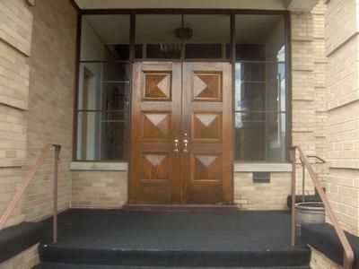 Brick building entrance with double wooden doors, glass side panels, and metal handrails up a short step.