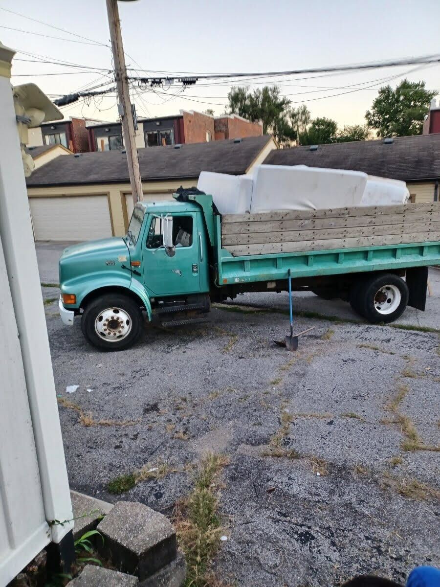 Teal dump truck carrying bricks parked in a lot, with buildings and power lines in the background.