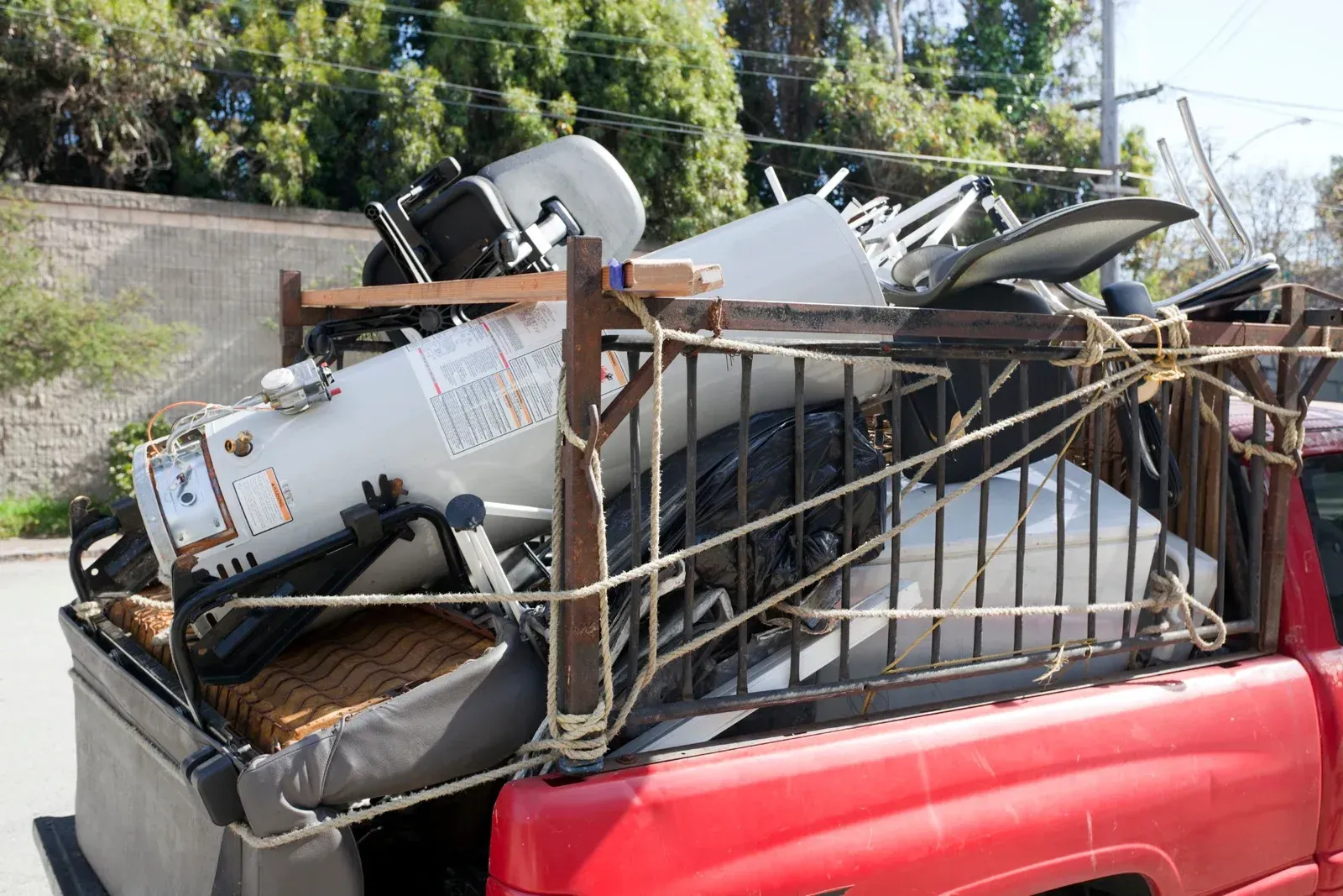 Red truck bed loaded with scrap metal and a water heater, secured with ropes.