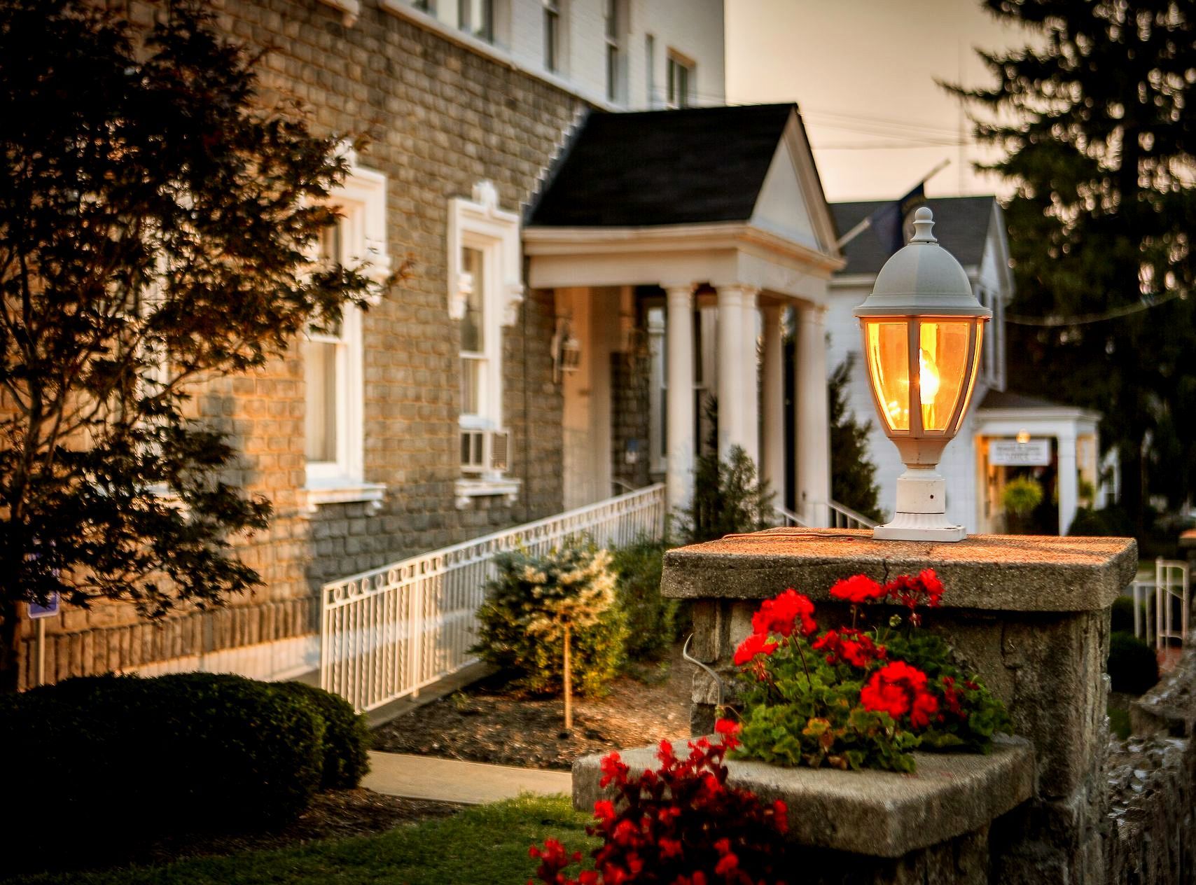 The Bentley House at dusk, with a lit lamp post and red flowers