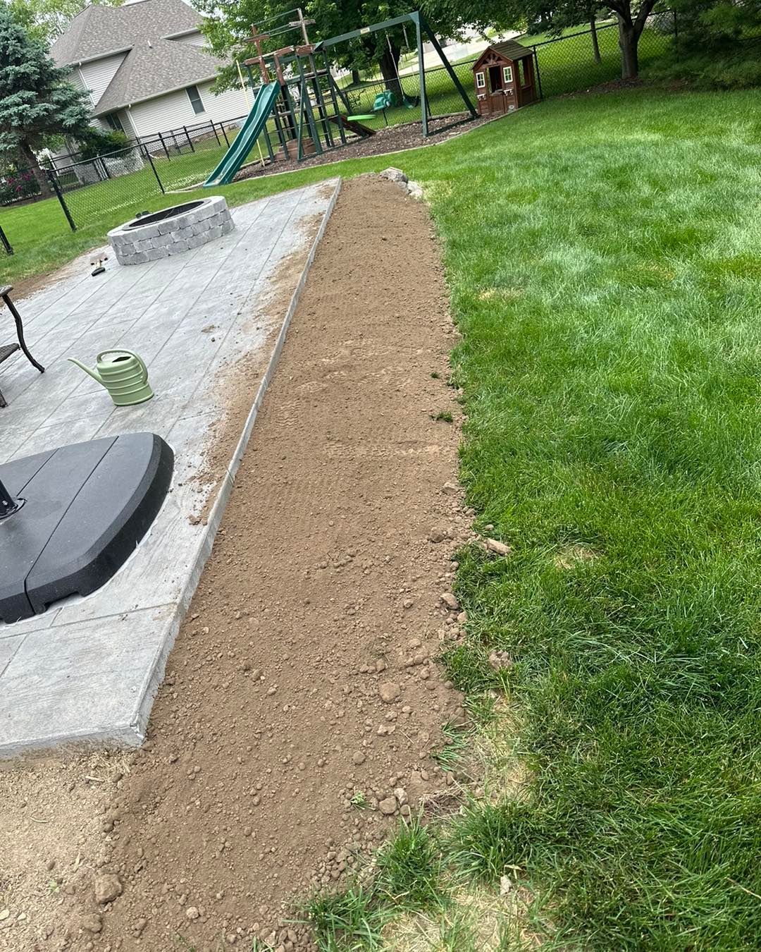A dirt strip borders a patio and green lawn in a backyard; a playground and house are in the background.