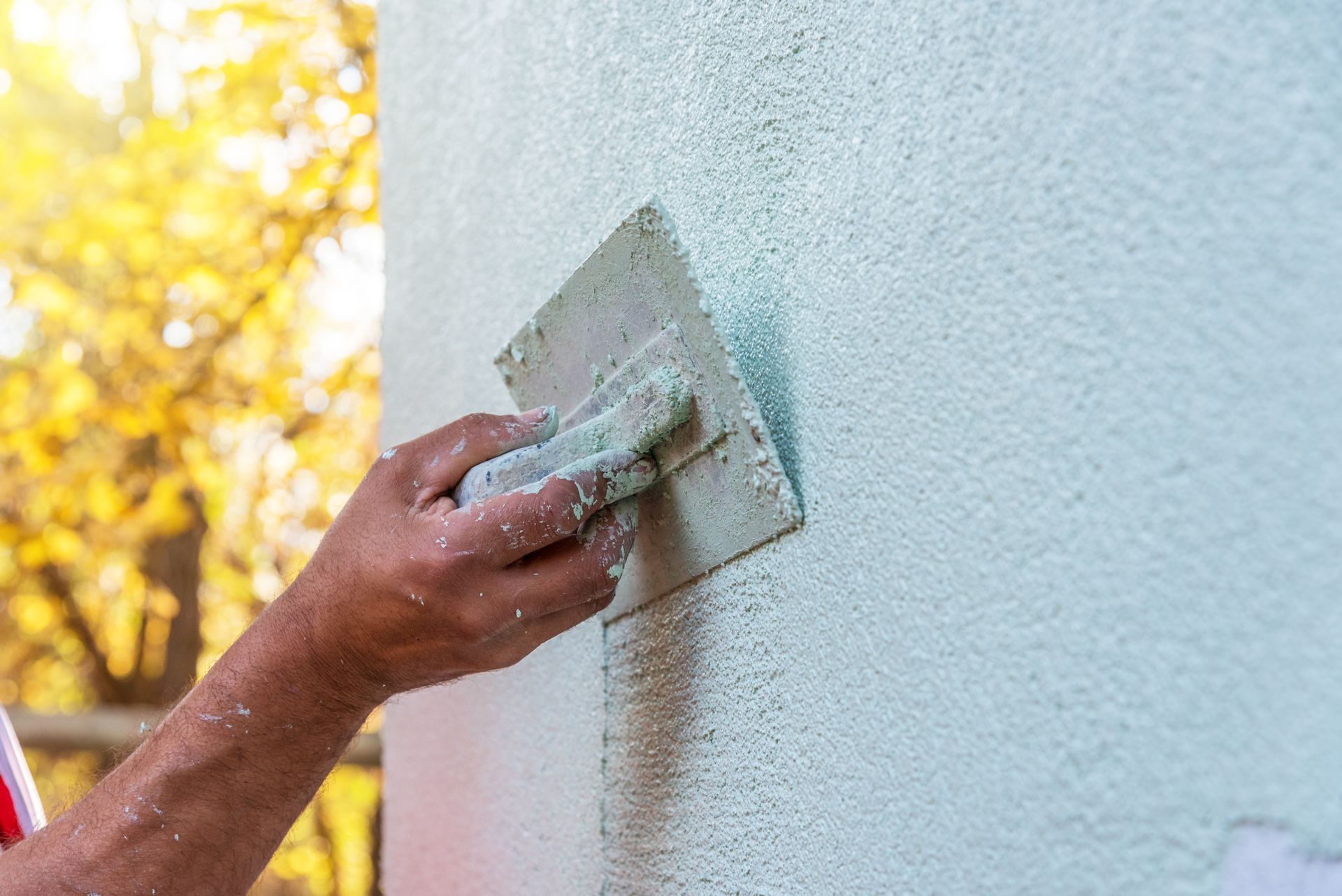 Eine Hand trägt mit einer Kelle hellblauen Putz auf eine strukturierte Außenwand auf.