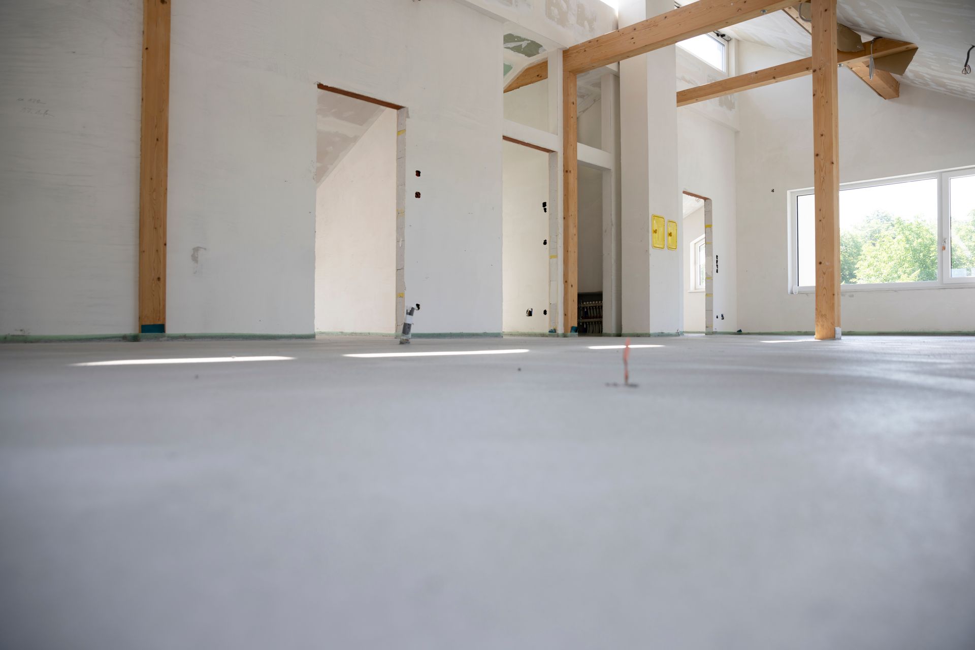 Ein heller, leerer, im Bau befindlicher Raum mit weißen Wänden, hölzernen Stützbalken und einem glatten, grauen Betonboden.
