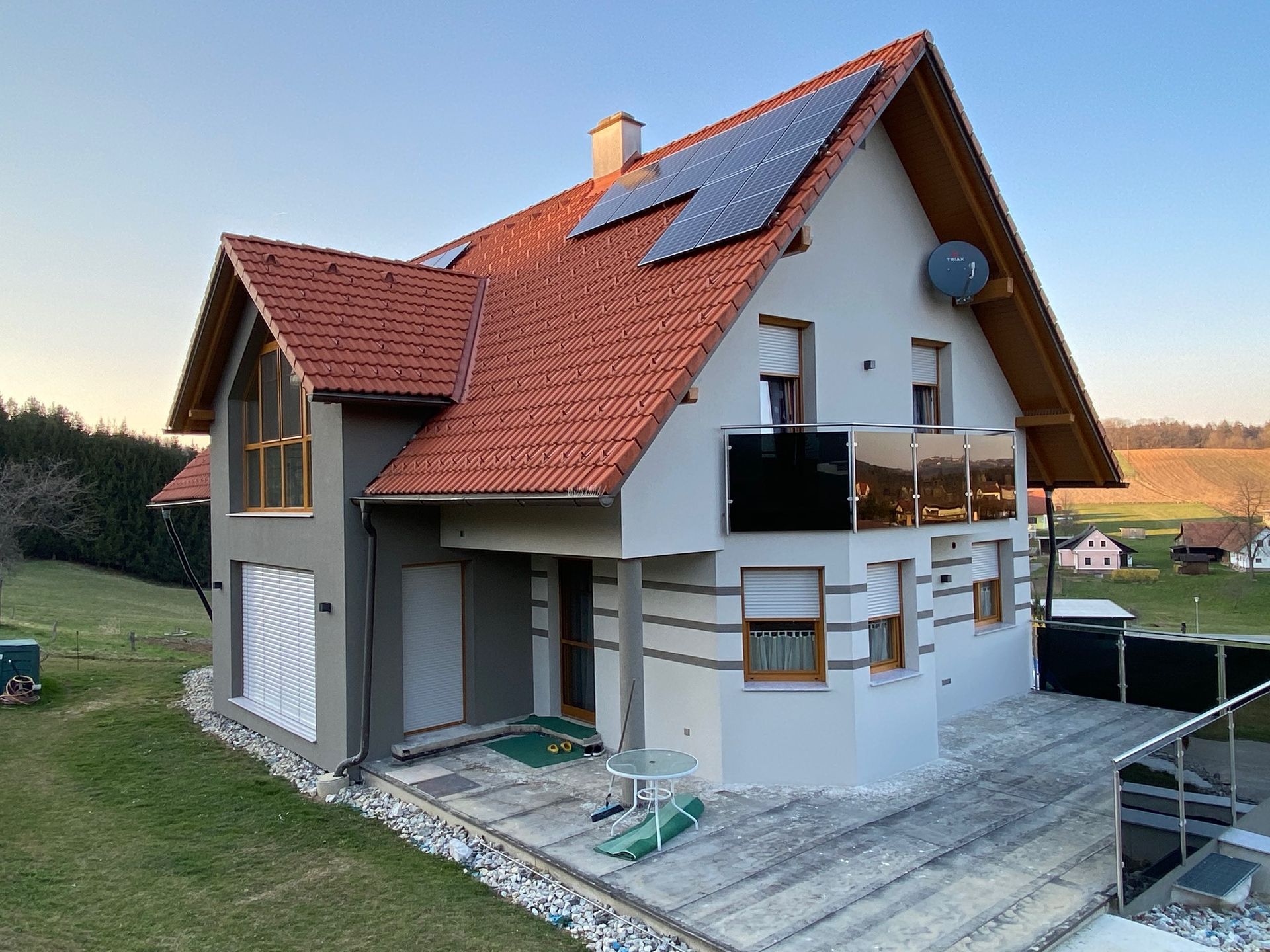 Ein helles, zweistöckiges Haus mit rotem Ziegeldach, Solarpaneelen, Balkon und Steinterrasse in ländlicher Lage.