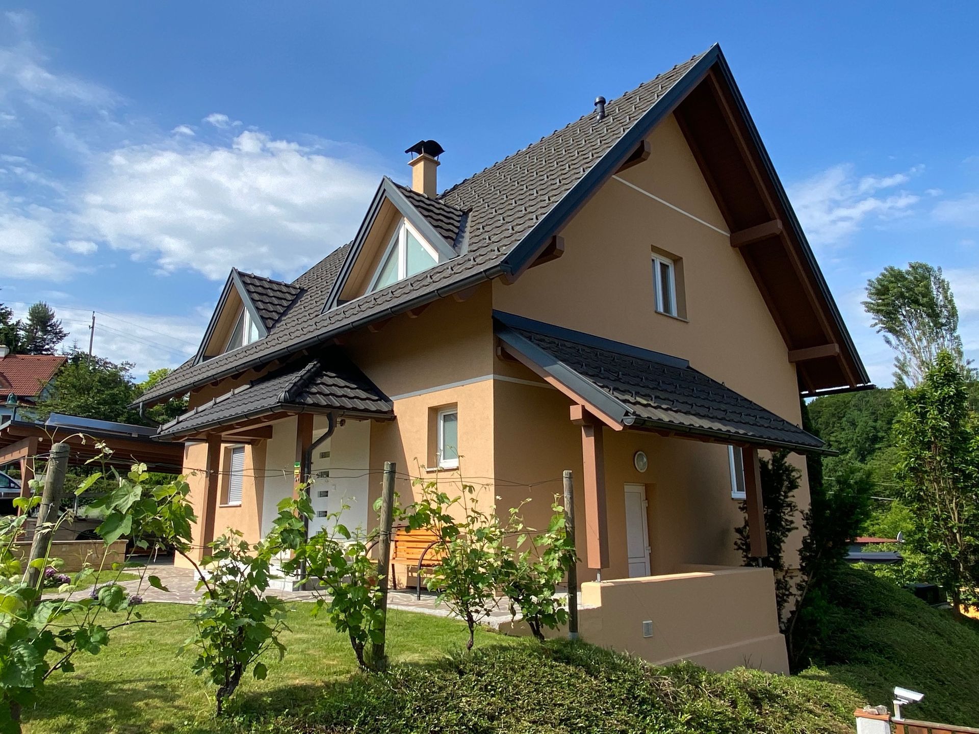 Ein zweistöckiges, hellbraunes Haus mit dunklem Giebeldach und Dachgauben, umgeben von grünen Weinreben