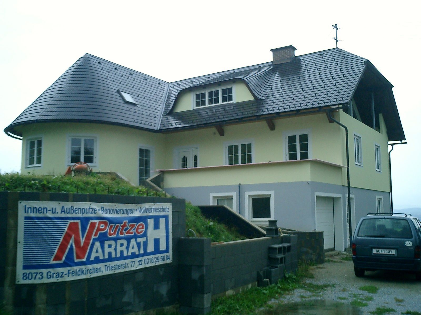 Ein zweistöckiges hellgelbes Haus mit dunklem Dach und grauem Keller, im Vordergrund ein Schild einer Baufirma.