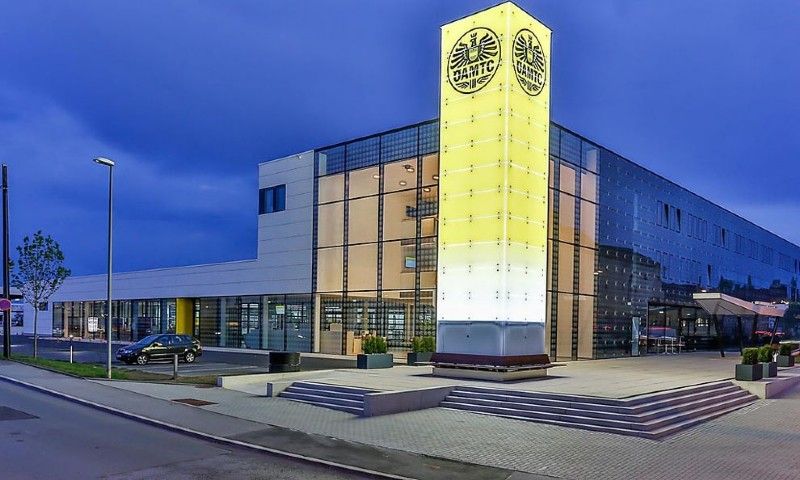 Ein modernes Gebäude in der Abenddämmerung mit einem hohen, beleuchteten, gelben Eckturm, der ein kreisförmiges Logo trägt.