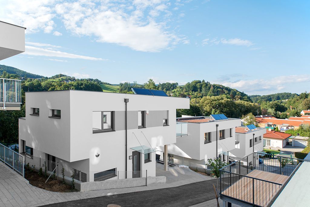 Moderne, mehrstöckige weiße Wohnhäuser mit Balkonen vor der Kulisse grüner Hügel und blauem Himmel.