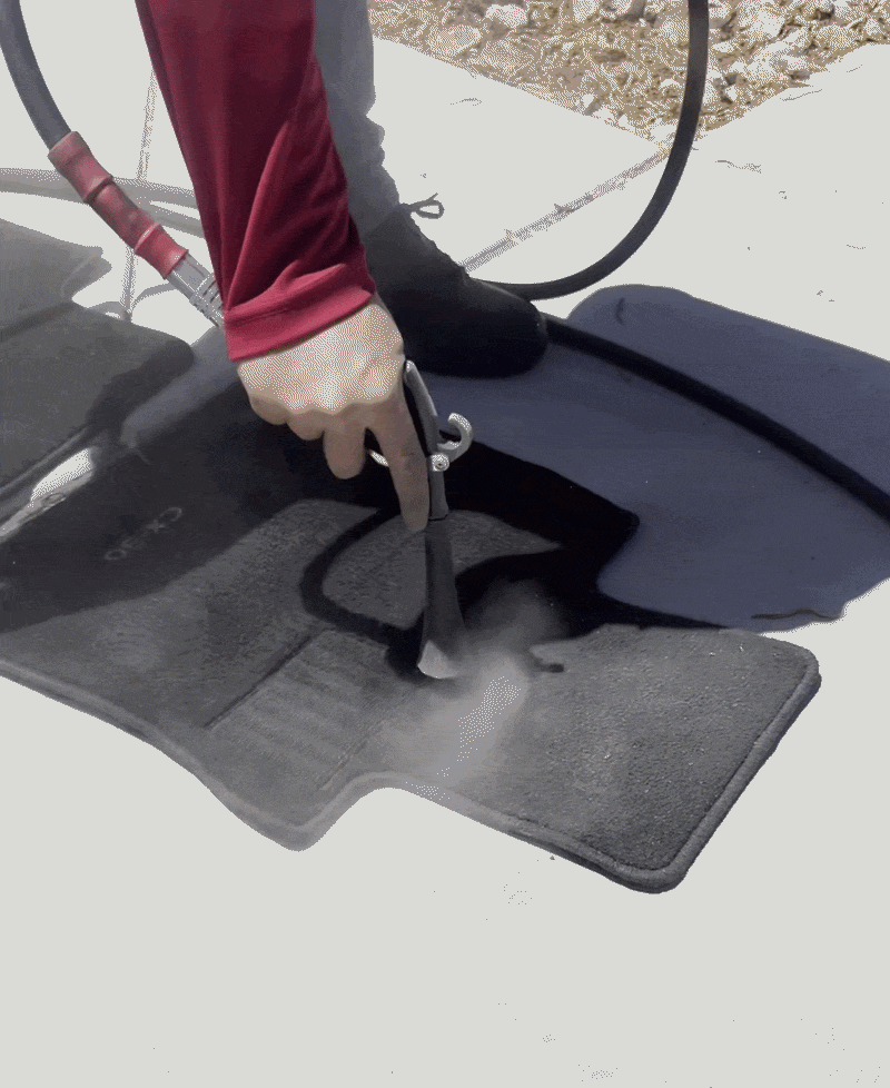 Person using a hose to wash a dark outdoor mat on concrete pavement.