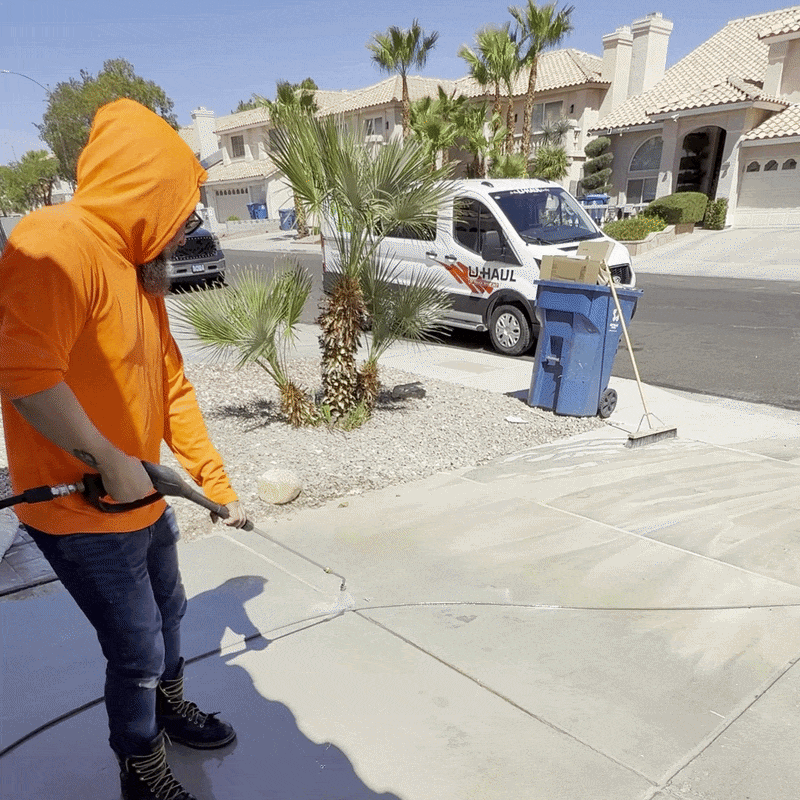 Worker in orange hoodie pressure washing a sidewalk on a sunny residential street.