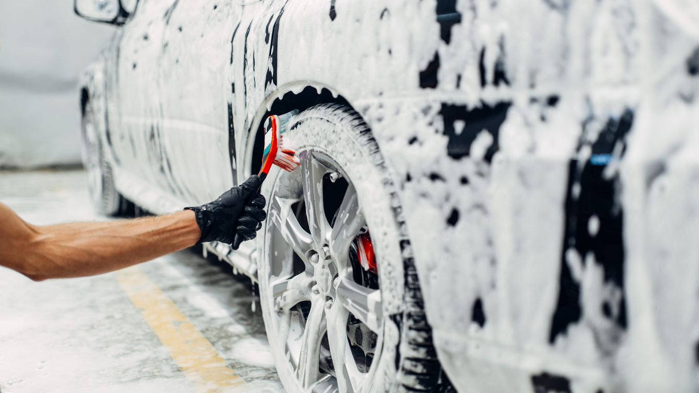 Person washing a white car wheel with soap suds and a brush in a car wash