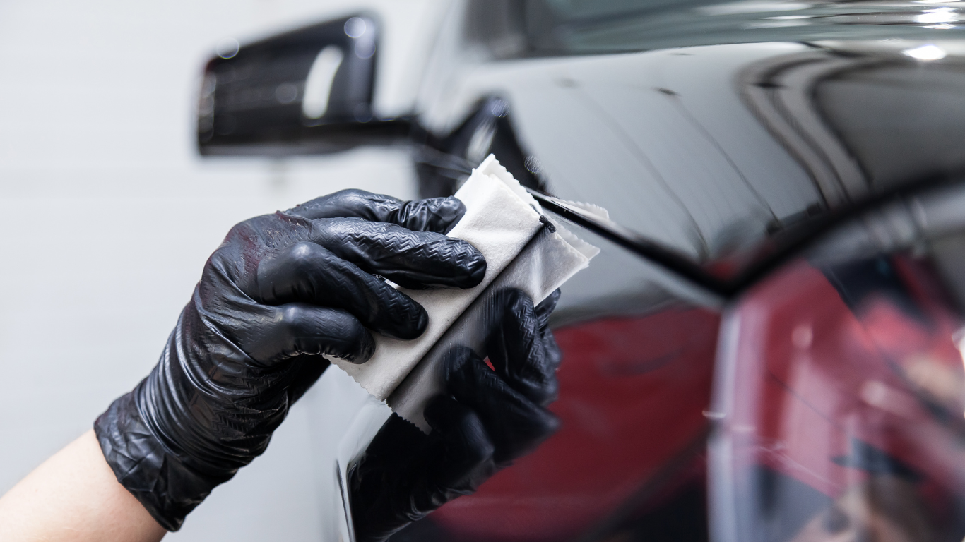 Gloved hand wiping a black car’s rear panel with a white cloth during detailing