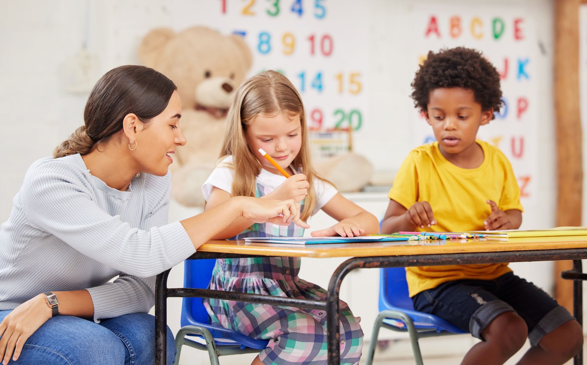 Teacher helping two children at a classroom table, with colorful numbers and letters on the wall