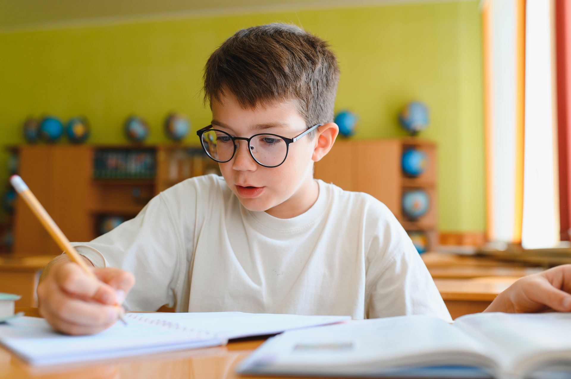 Boy wearing glasses writing in a notebook at a desk in a bright classroom