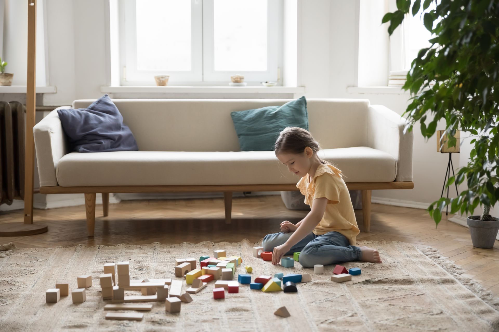 Child playing with wooden blocks on a rug in a bright living room with a white sofa