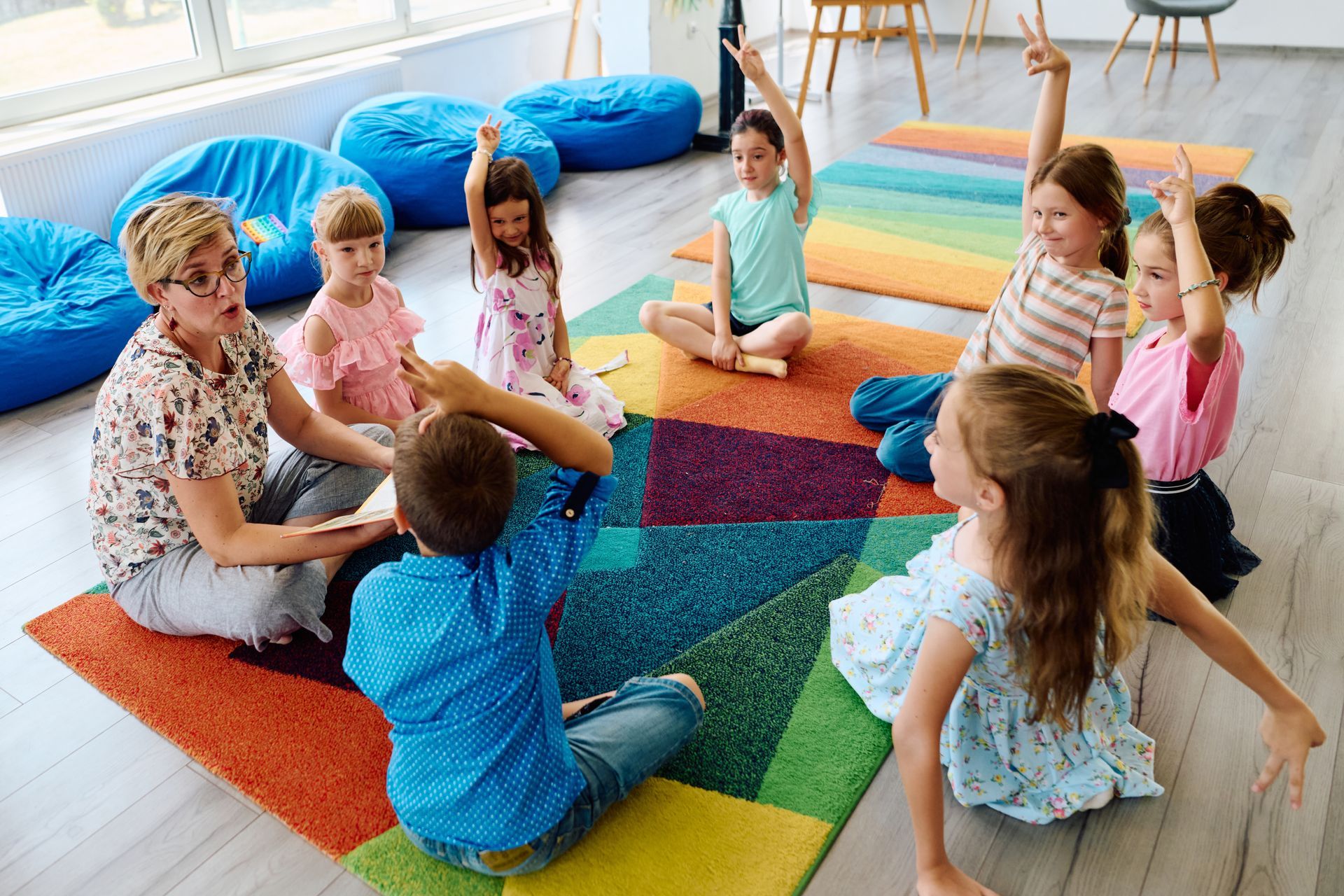 Children sitting on colorful mats in a bright classroom, facing a seated teacher during circle time