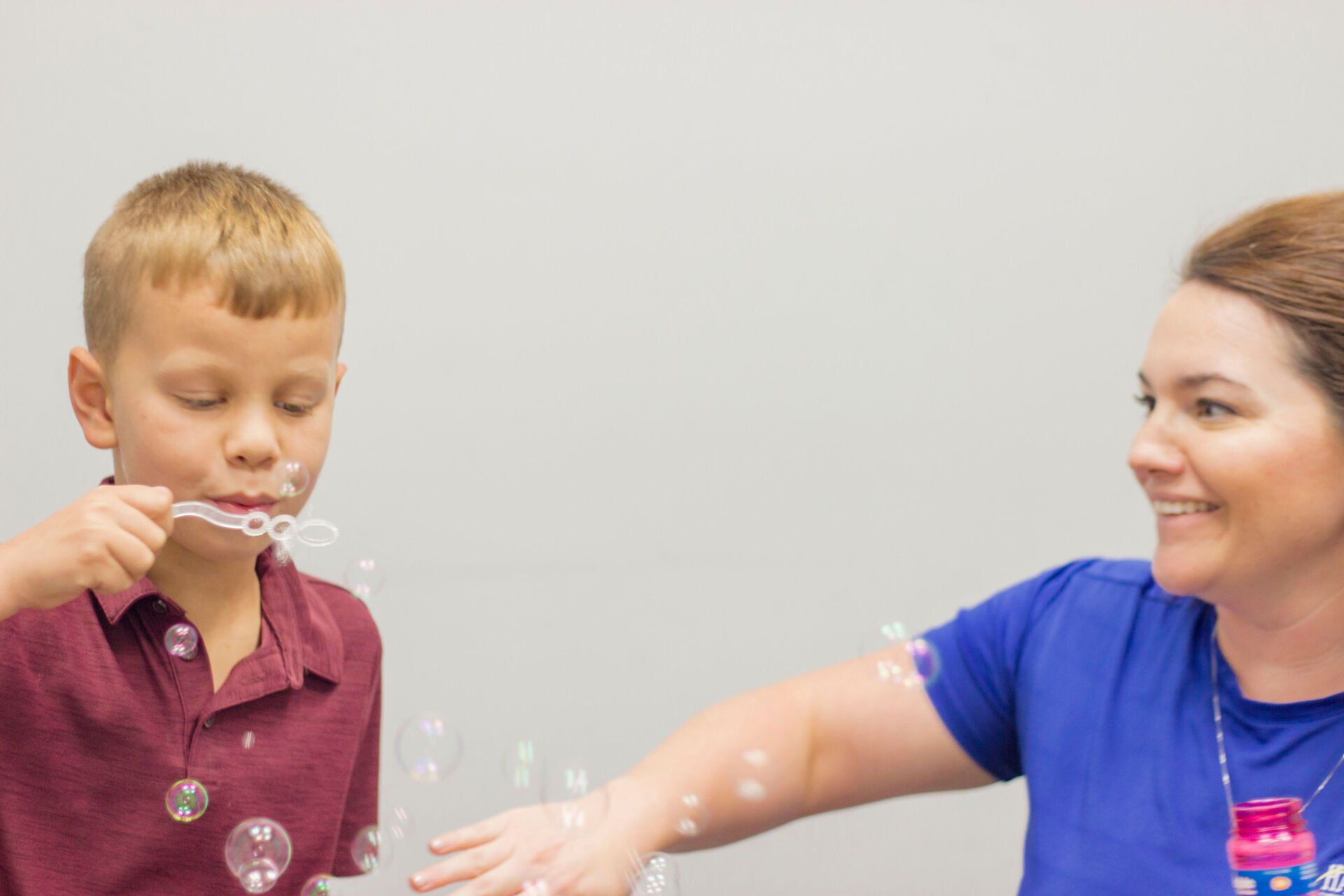 Child blowing bubbles while a woman smiles and reaches out in a bright room.