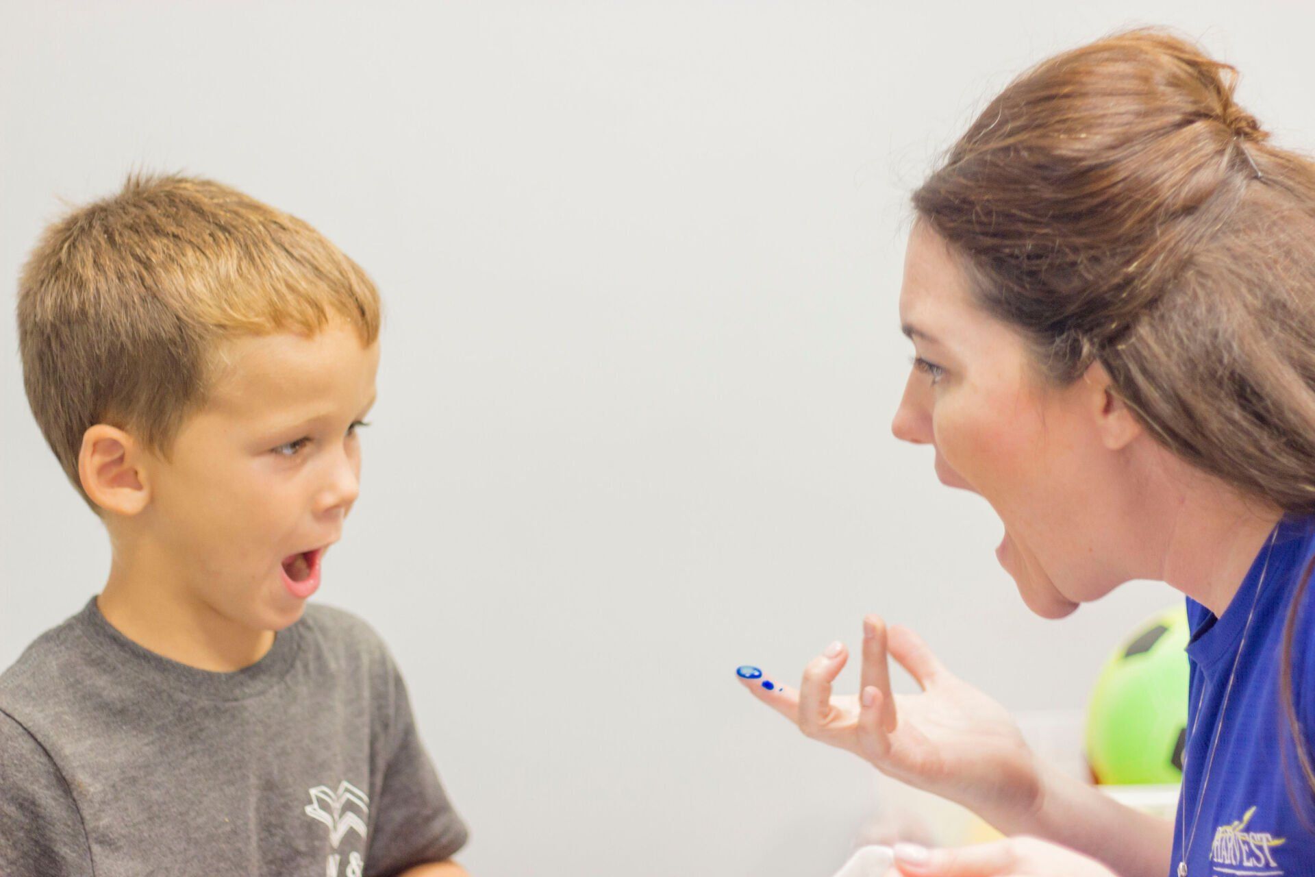 Adult and child facing each other with open mouths, appearing to practice speech in a bright room