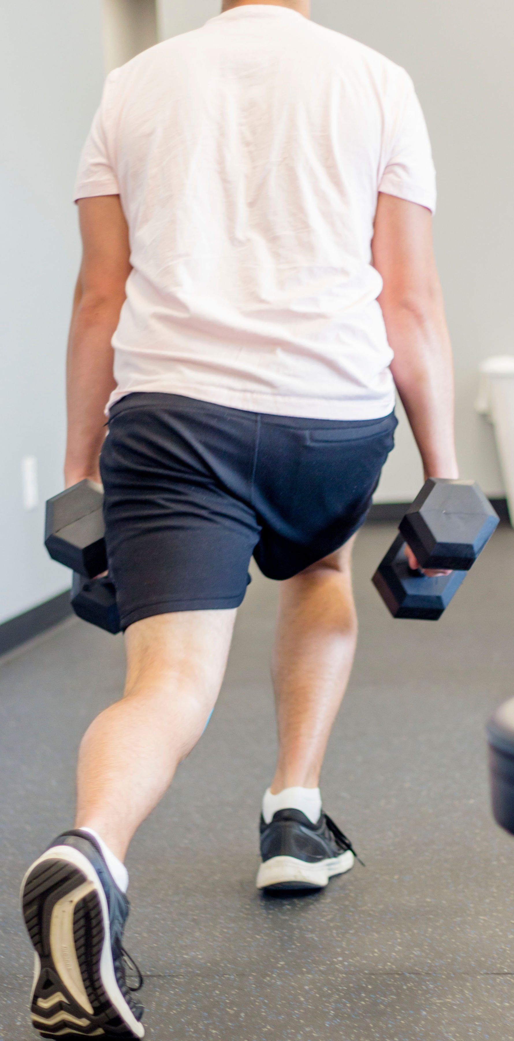 Person doing a dumbbell walking lunge in a gym, wearing a white shirt and dark shorts.