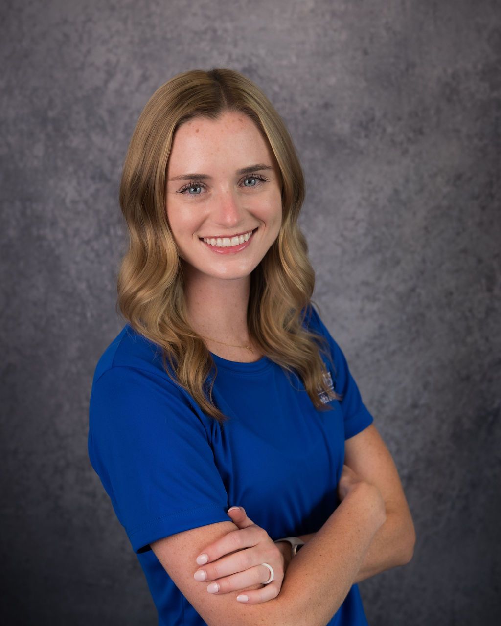 Smiling woman in a blue shirt with arms crossed against a gray studio backdrop