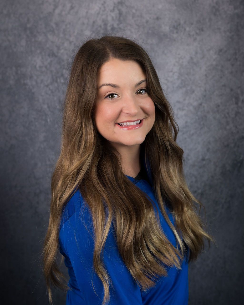 Smiling woman with long brown hair in a blue top against a gray backdrop