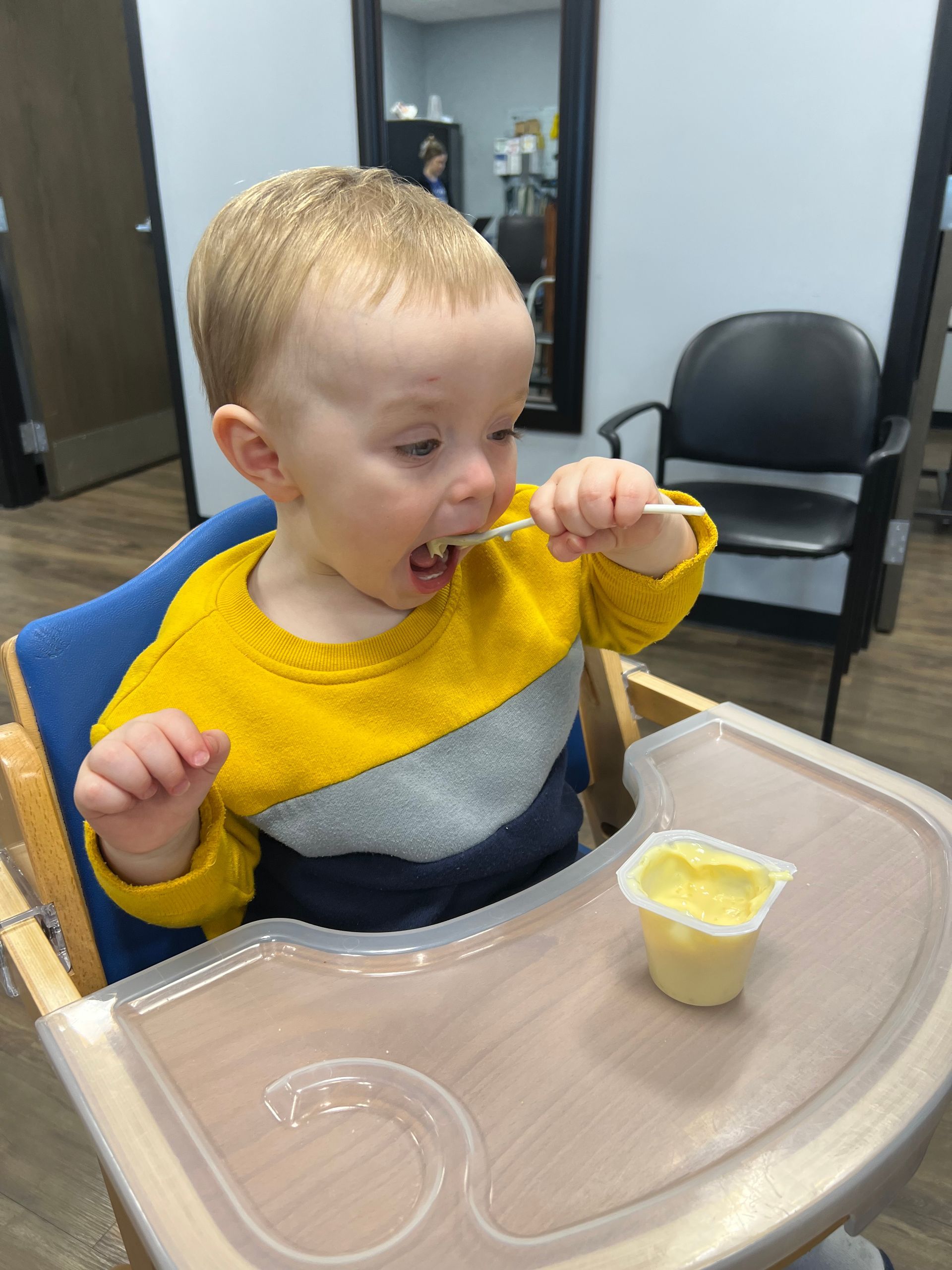 Child eating with a spoon at a tray table, wearing a yellow and gray sweater, with a small cup of food nearby