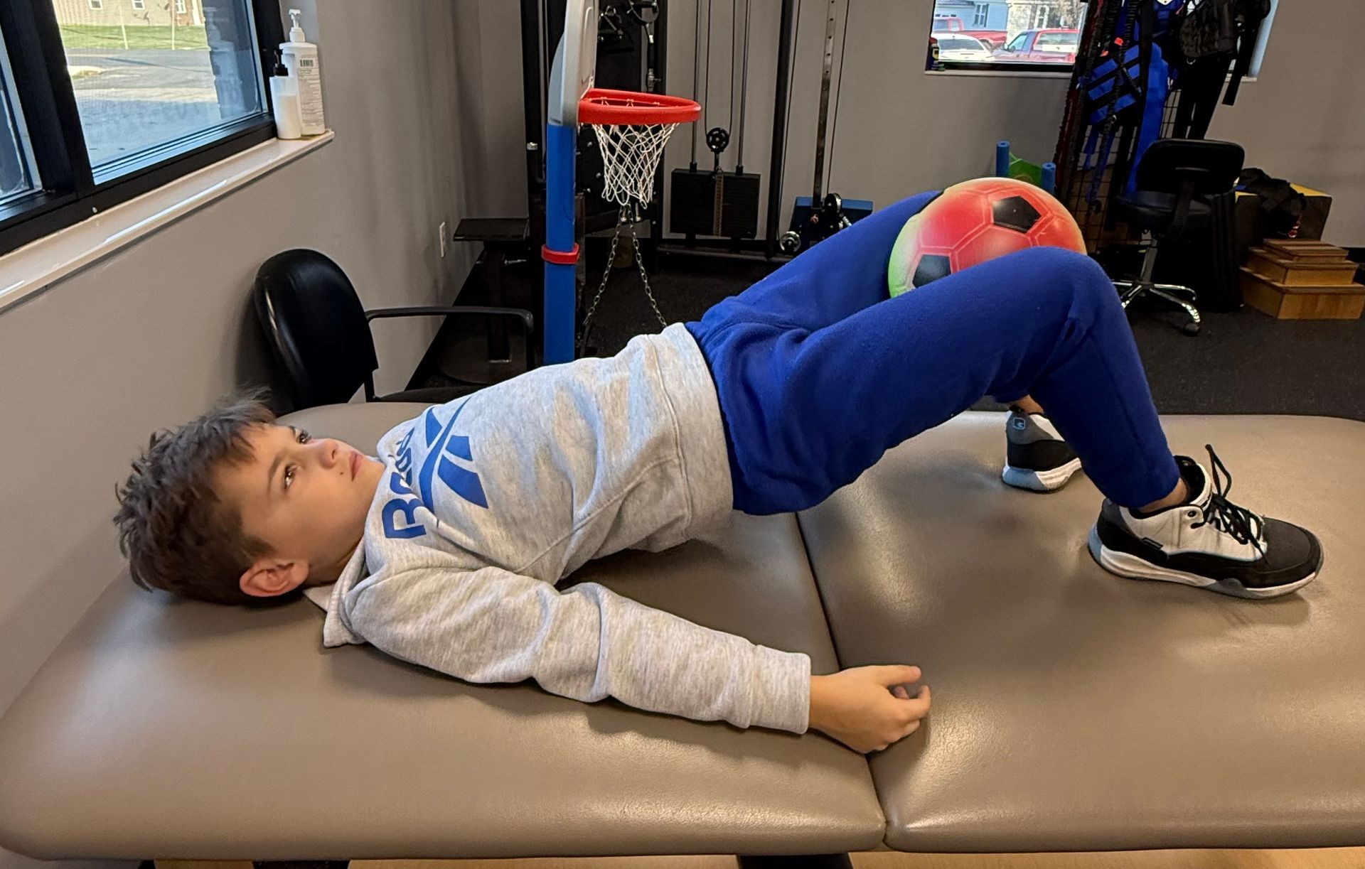 Child doing a bridge exercise on a therapy table in a clinic, with a red ball nearby.