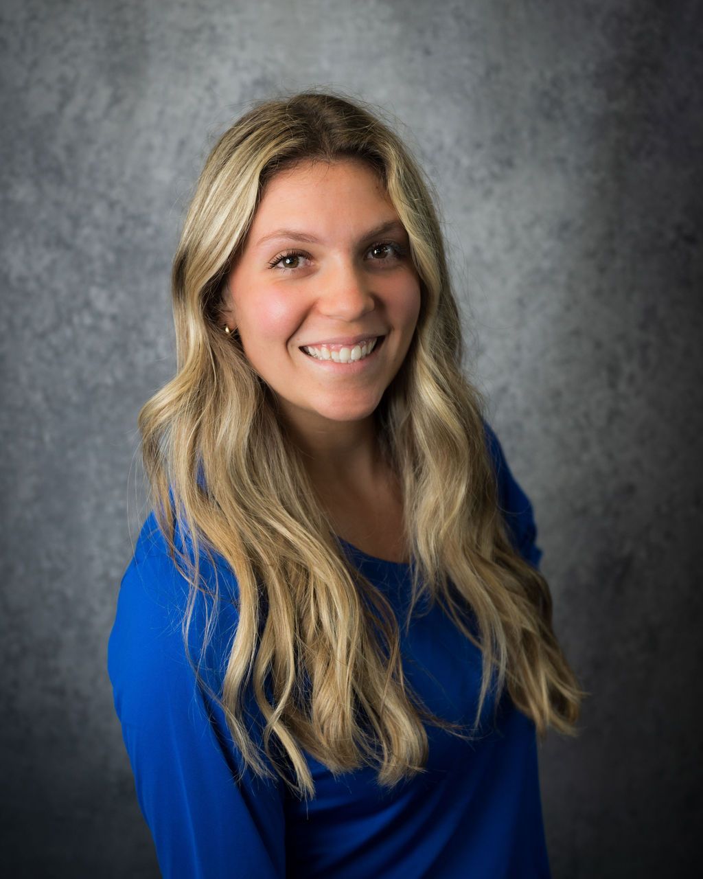 Smiling woman with long blonde hair wearing a blue top against a gray studio backdrop