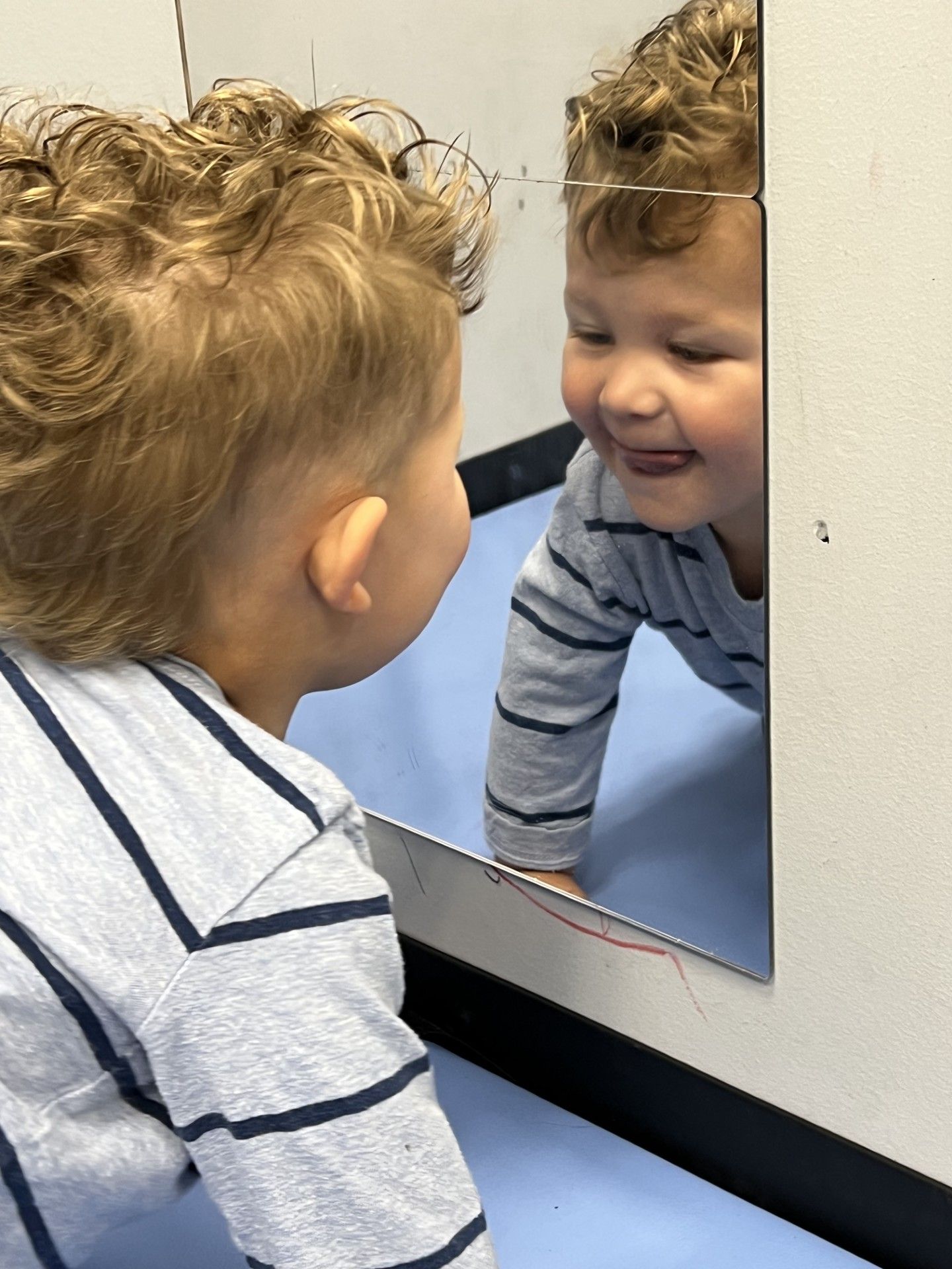Child smiling at their reflection in a mirror, wearing a striped shirt indoors.