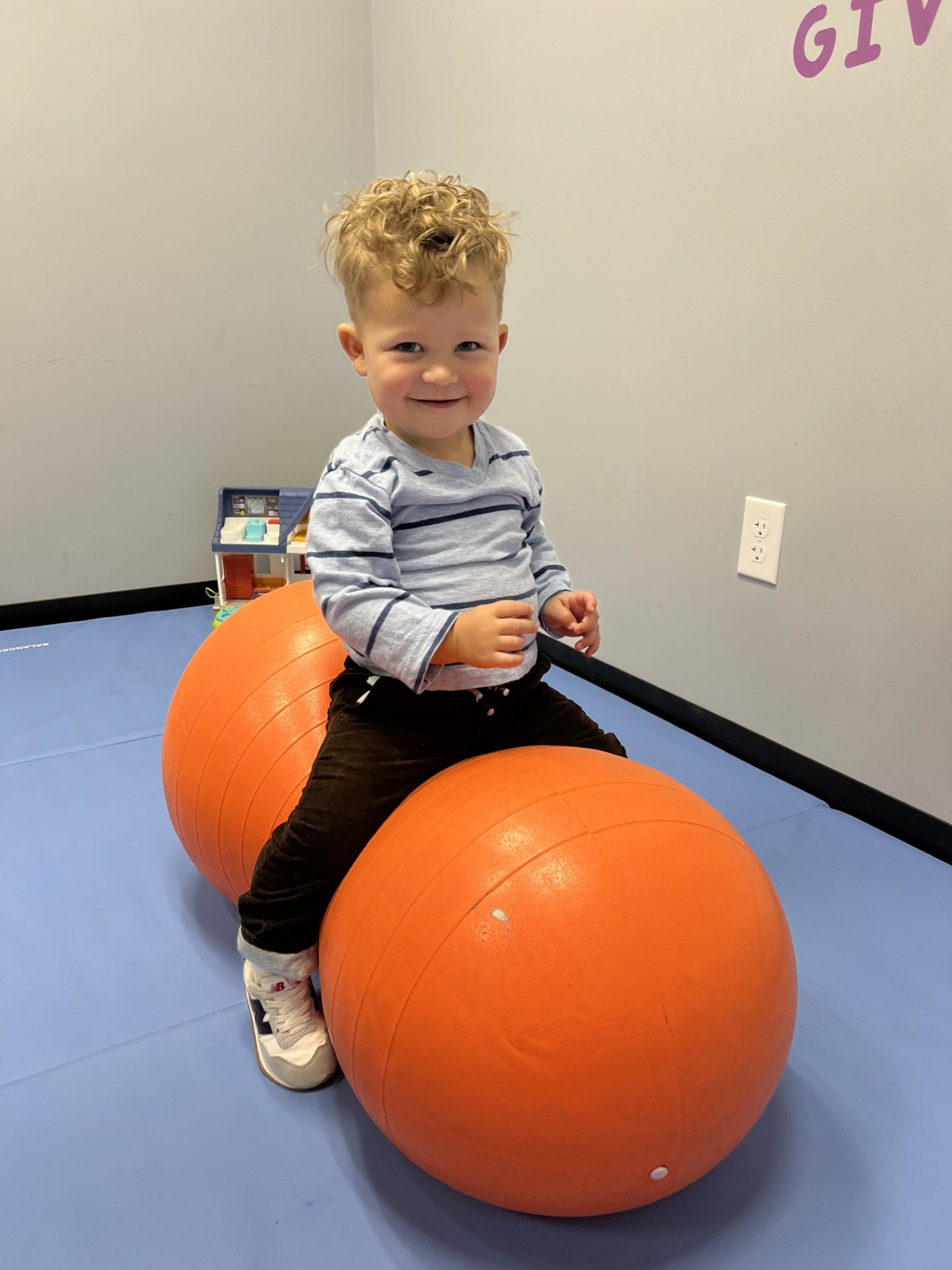 Child sitting on an orange exercise ball in a therapy room, smiling at the camera.