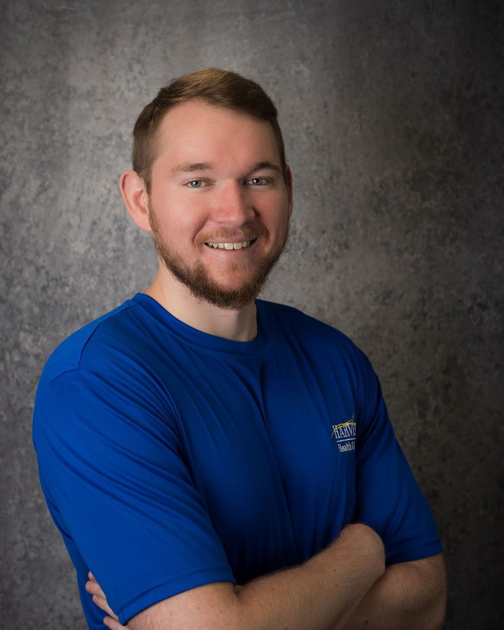 Smiling man in a blue shirt with arms crossed against a gray studio backdrop