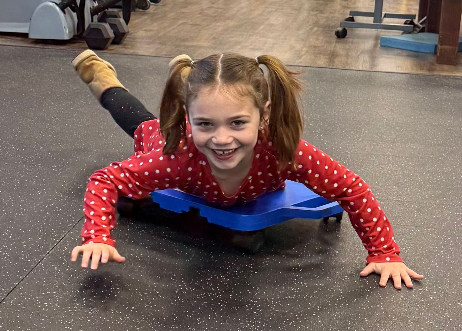 Child smiling on a blue balance board in a gym, wearing a red polka-dot shirt.