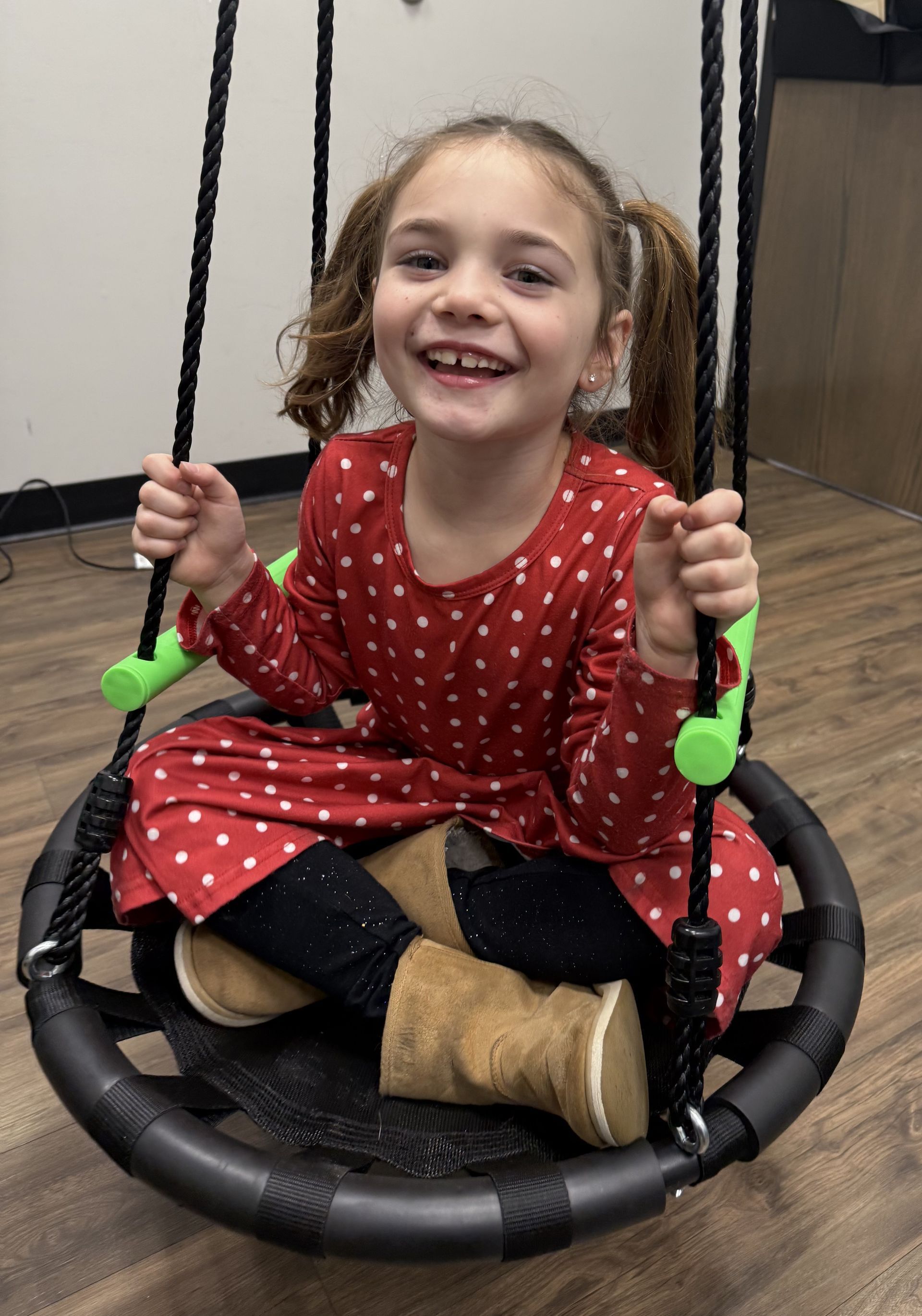 Smiling child sitting on a black circular swing, wearing a red dress and tan boots.