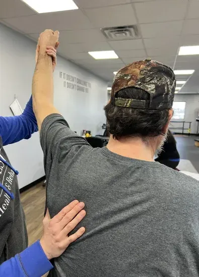 Person in camouflage cap is being assisted with an arm raised in a bright indoor room