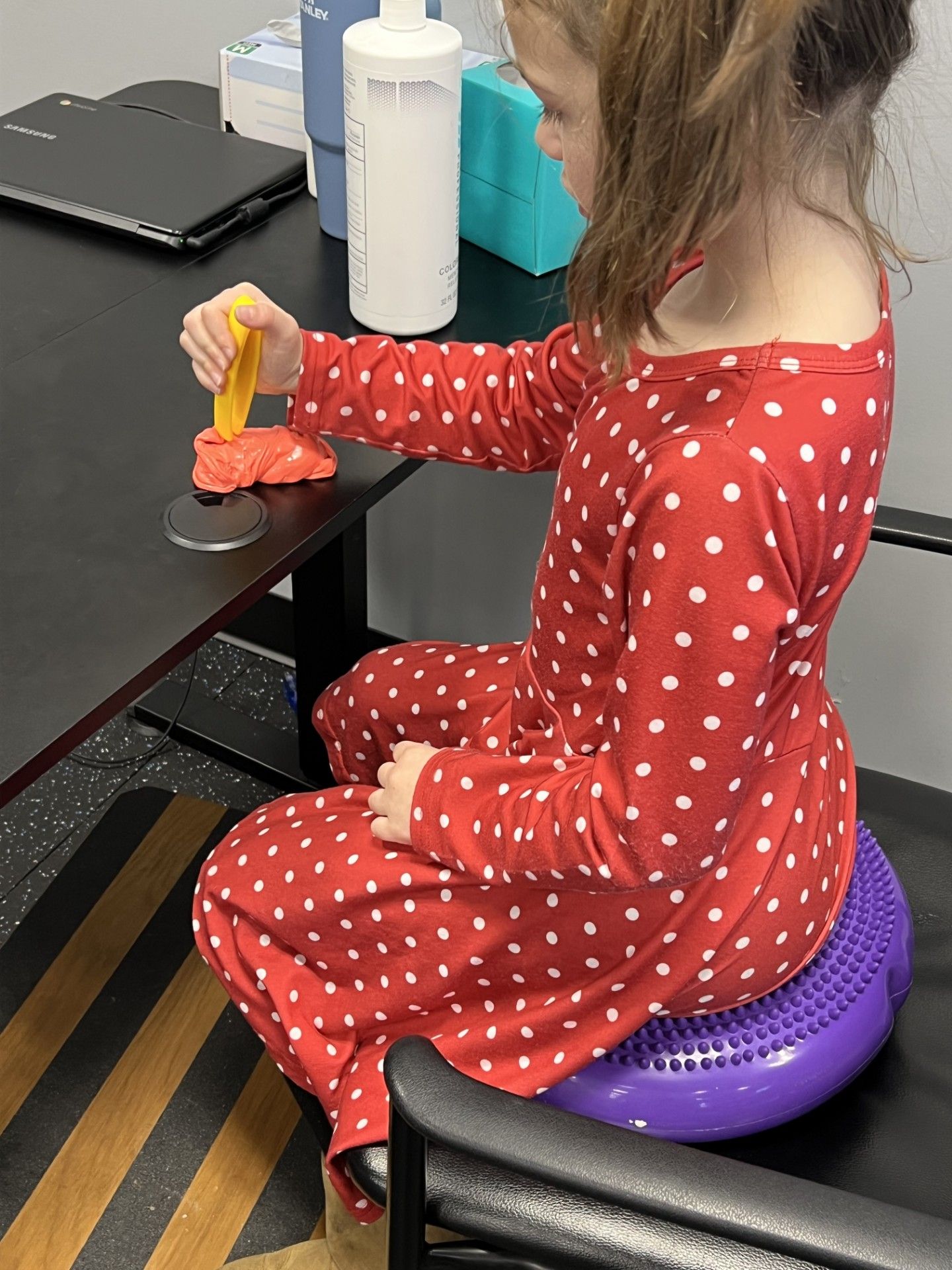 Child in red polka-dot pajamas using a toy hammer at a table, seated on a purple cushion.