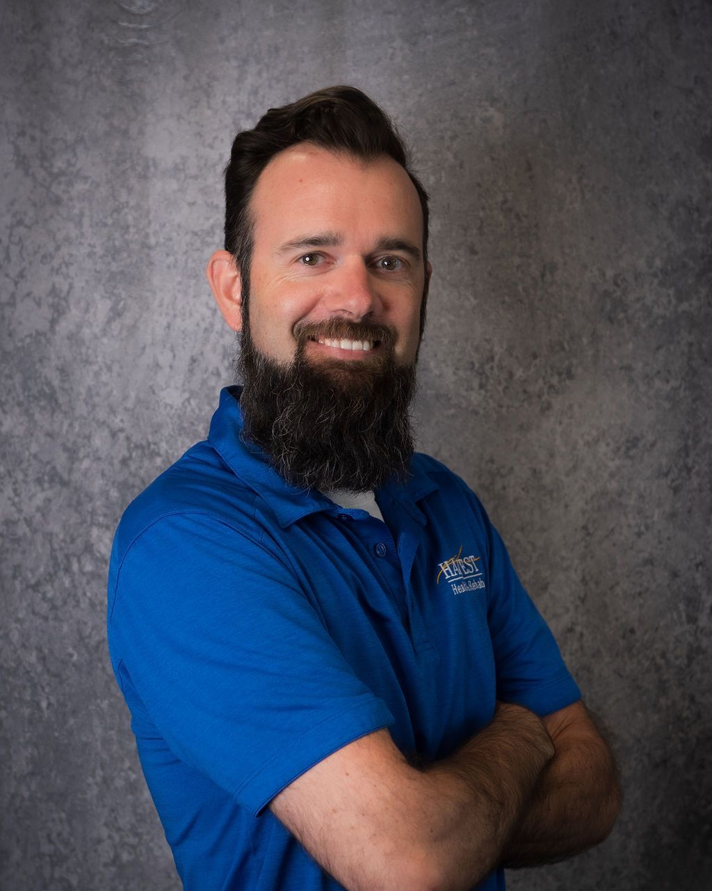 Smiling man in a blue polo shirt with arms crossed against a gray studio backdrop