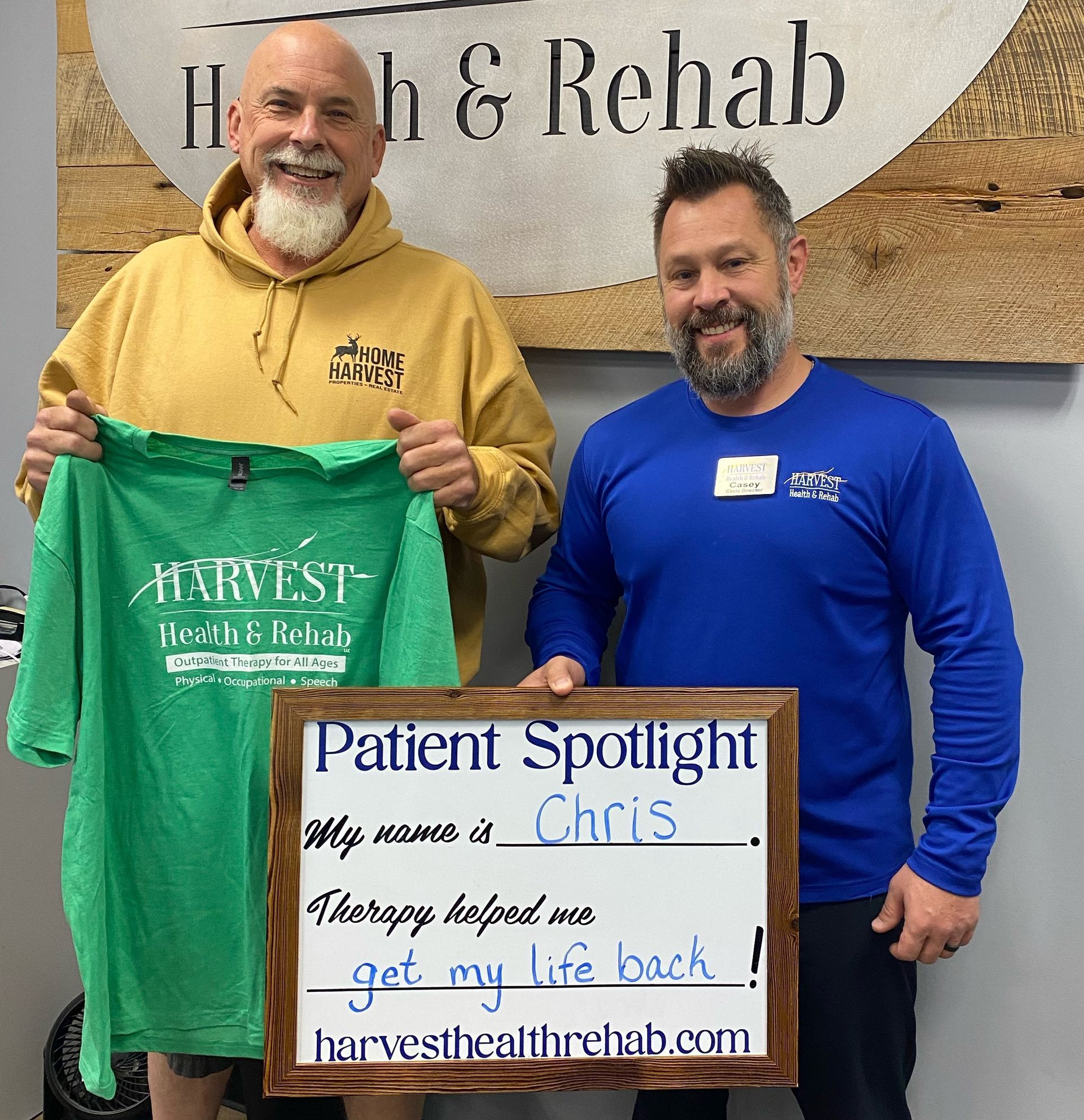 Two men holding a green shirt and a “Patient Spotlight” sign in a rehab office