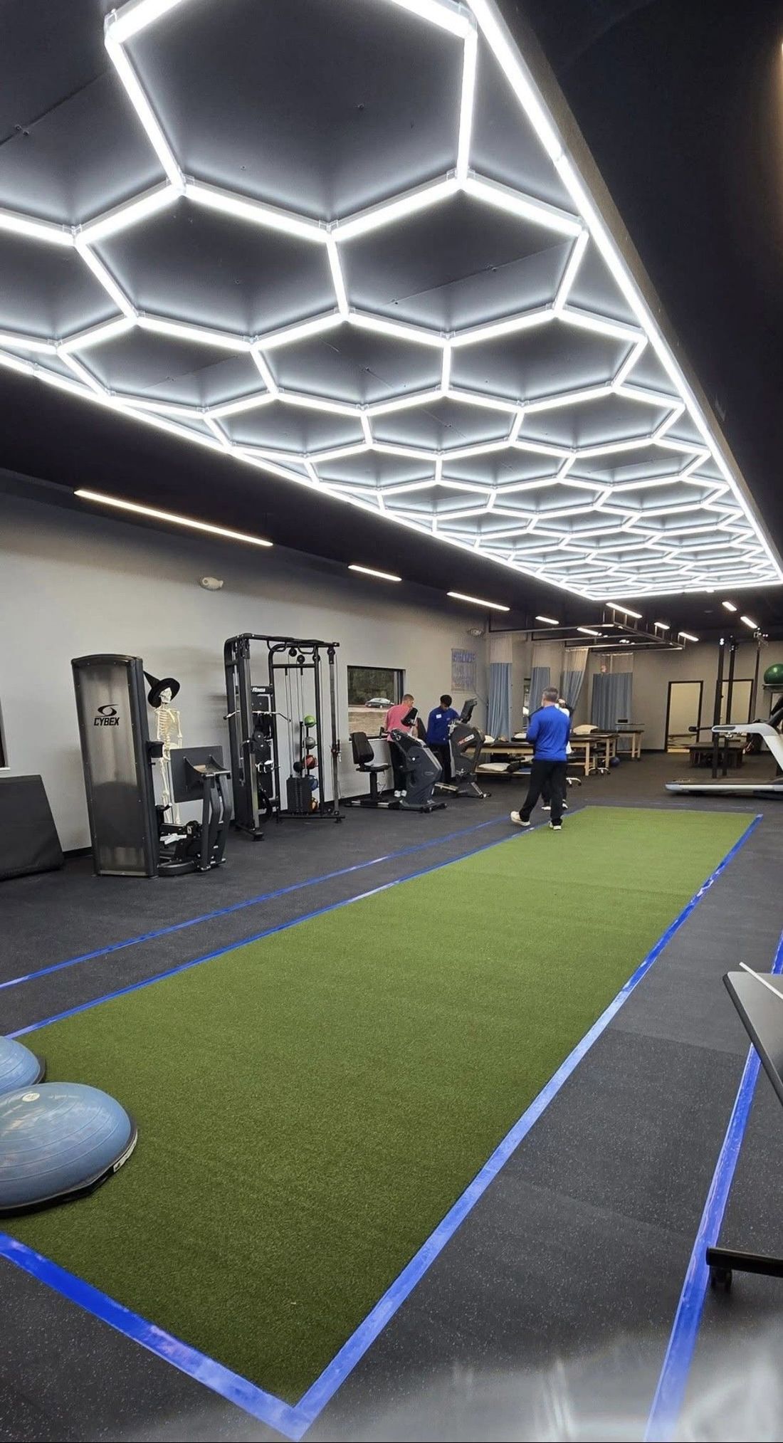 Indoor gym with hexagonal ceiling lights, green turf lane, and people training near exercise equipment.