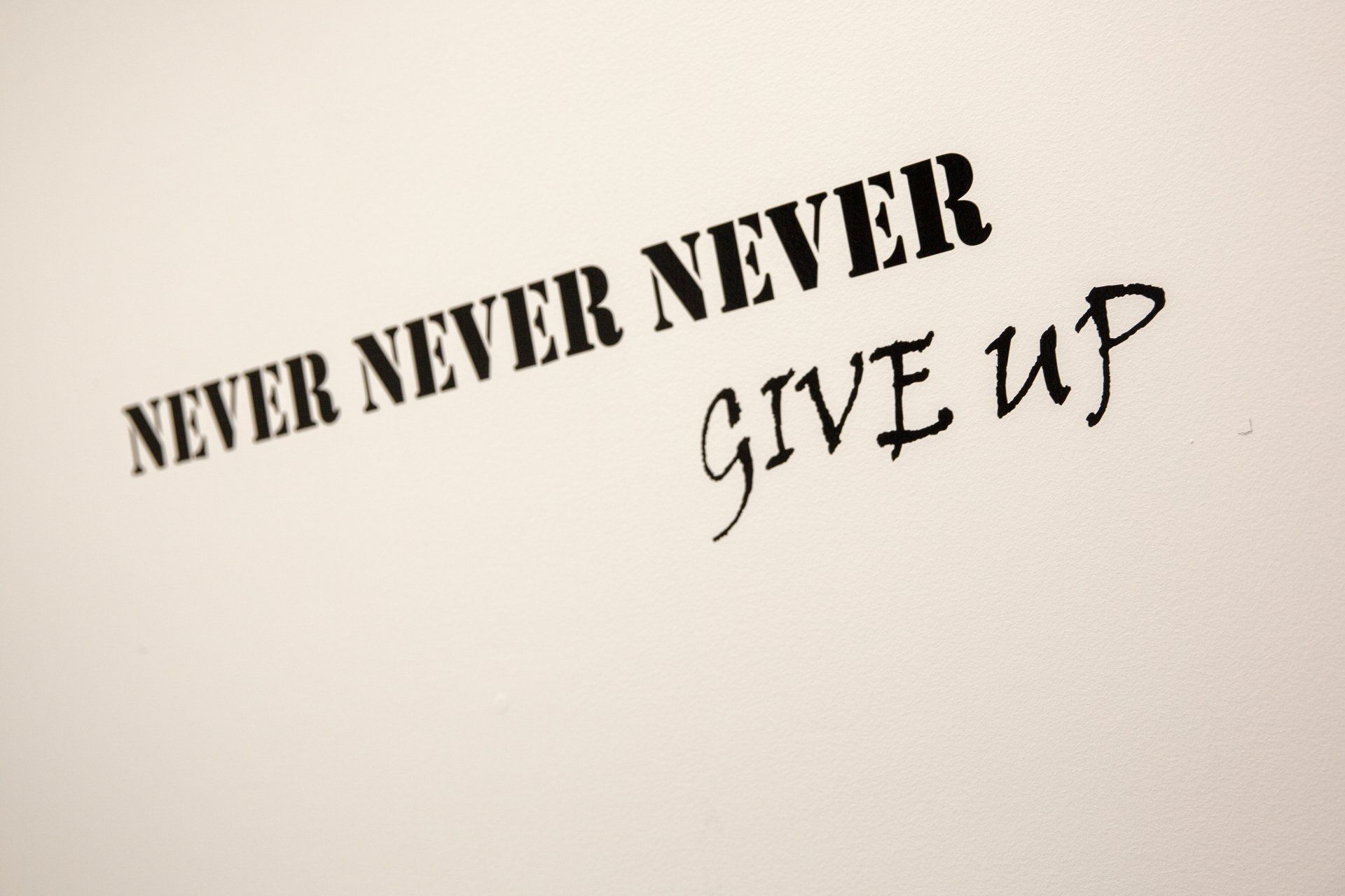 Inspirational wall text reading “Never Never Give Up” in black lettering on a light background