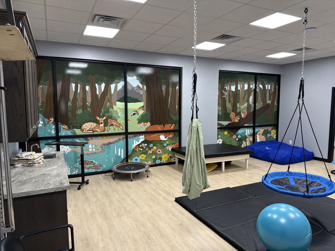 Indoor sensory room with swings, mats, and therapy equipment, featuring a forest mural wall.