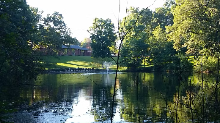 A calm lake reflects the surrounding trees and sky, with a fountain and a house in the distance.
