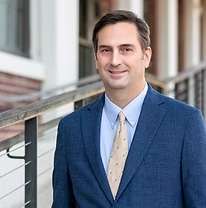 Man in blue suit, light blue shirt, patterned tie, smiling outdoors, near railing.