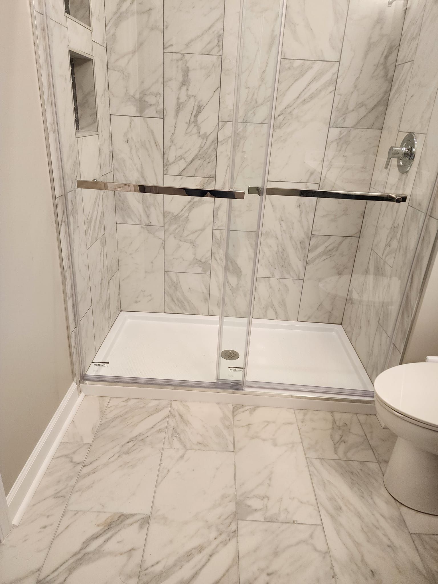 Marble-tiled shower with glass doors and a white base, next to a toilet.