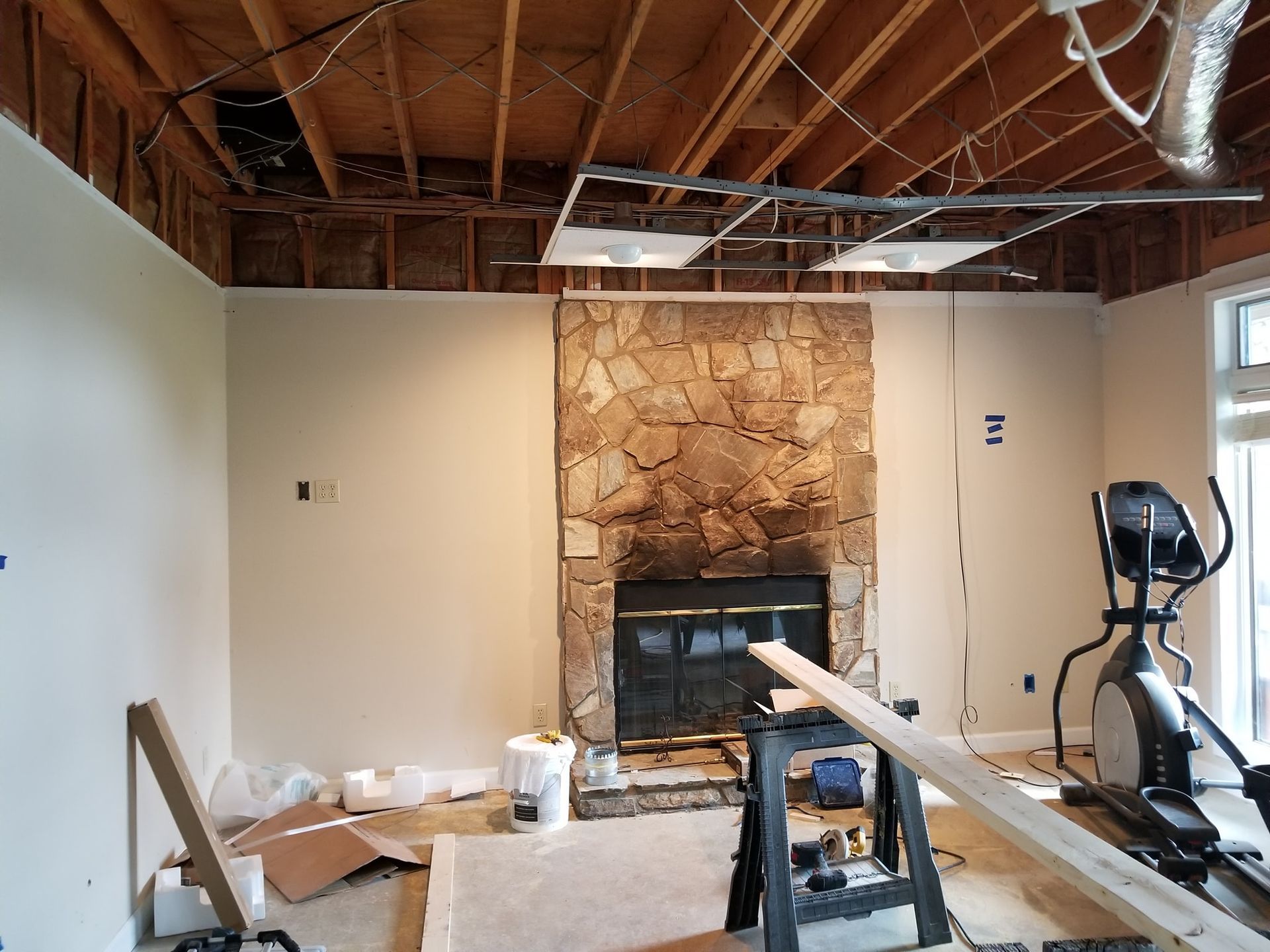 Room under renovation with stone fireplace, exposed ceiling beams, and exercise equipment.