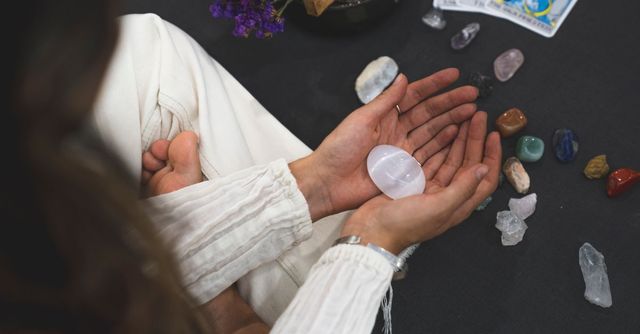 Unas manos con mangas blancas sostienen un cristal transparente sobre una mesa oscura con piedras de colores esparcidas