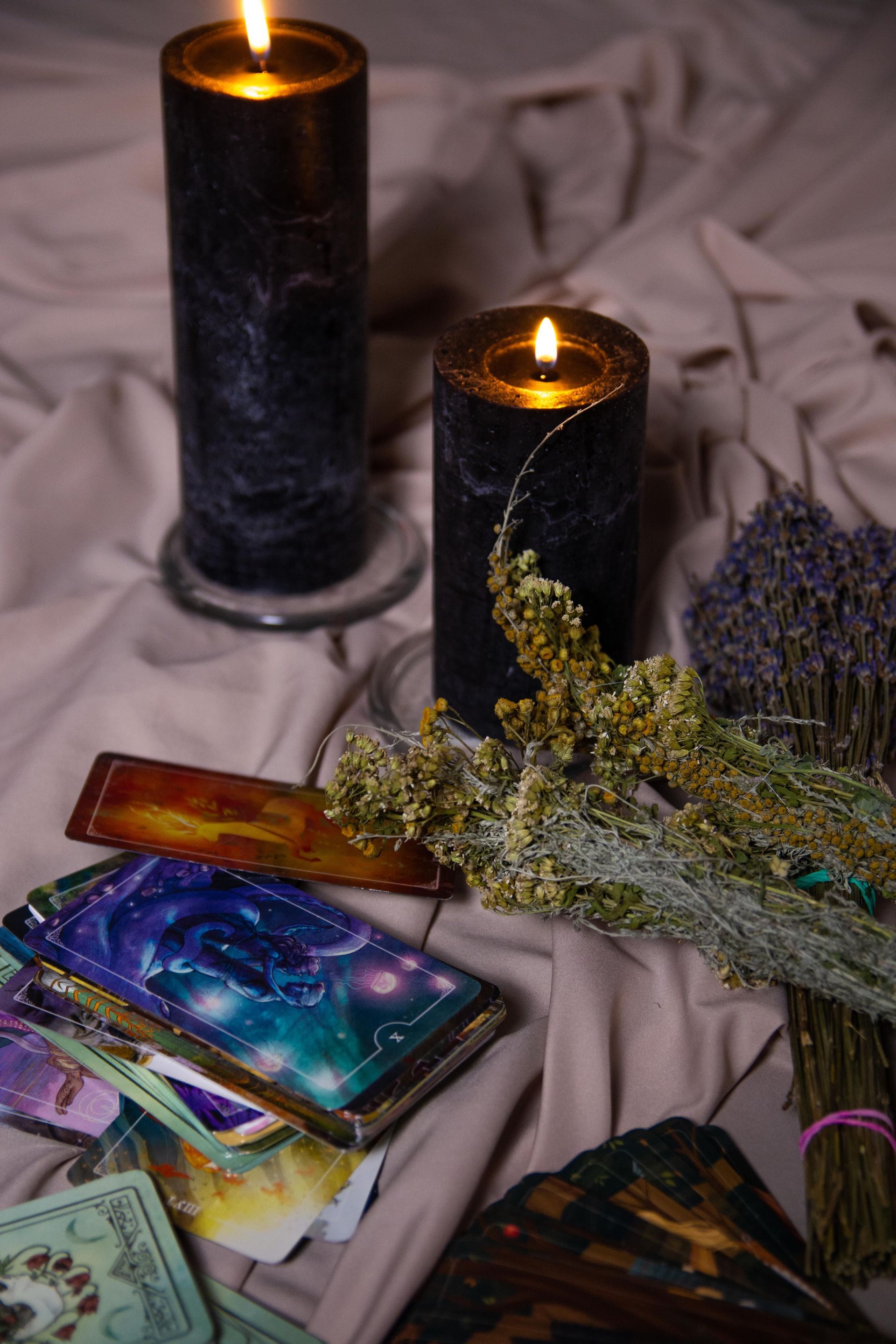 Dos velas negras junto a unas cartas del tarot y flores secas sobre una tela arrugada