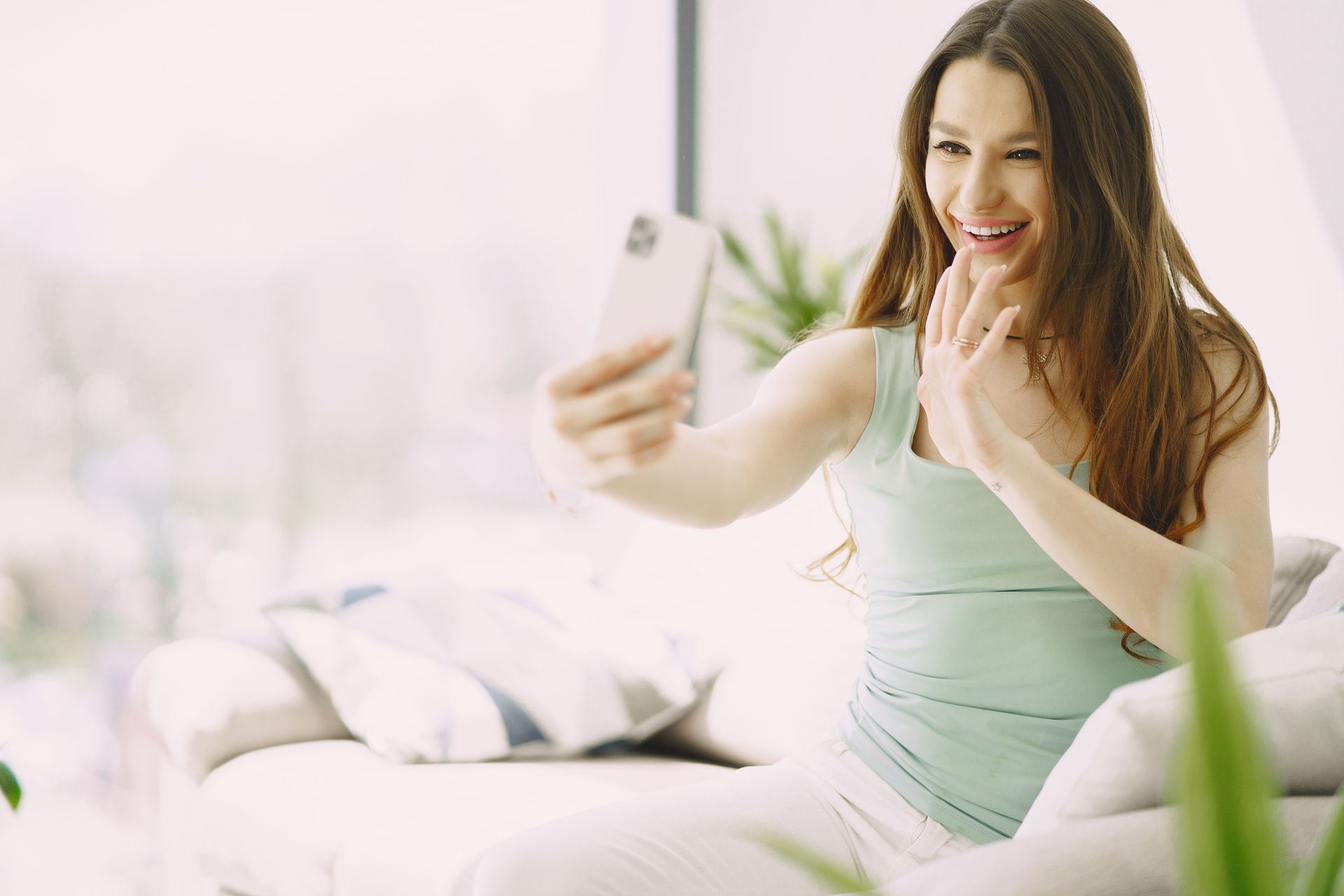 Una mujer se hace un selfie en la cama, en un dormitorio luminoso, sonríe y saluda con la mano.