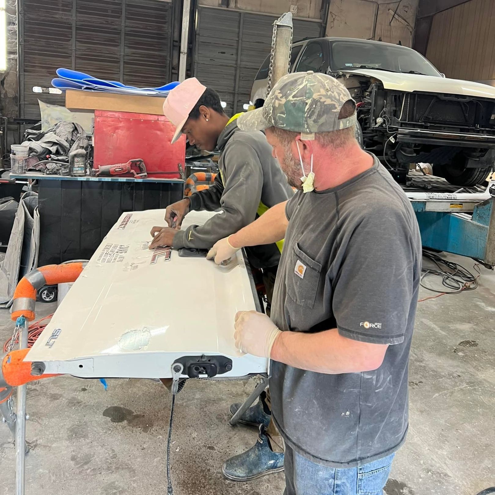 Two people work on a white vehicle panel in a garage, one pointing at the surface while the other holds a tool.
