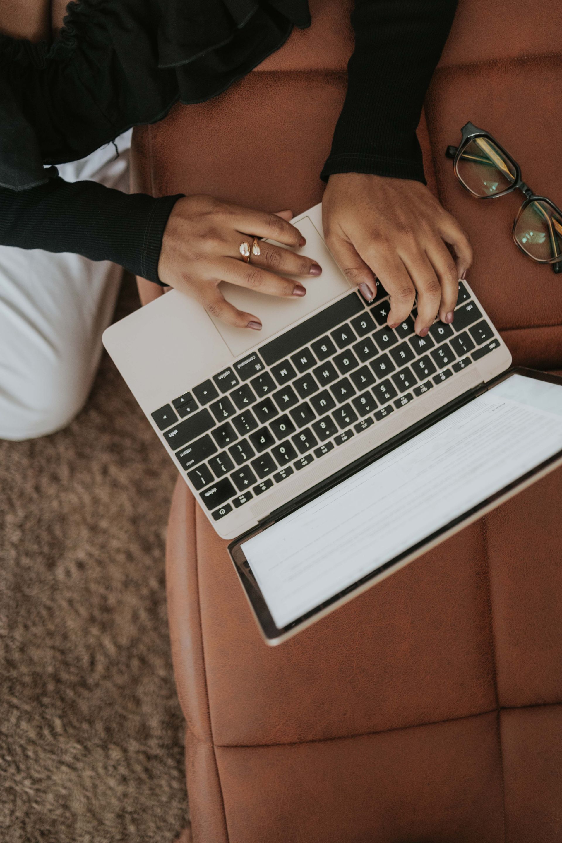 Hands typing on a laptop on a brown couch beside a pair of glasses.